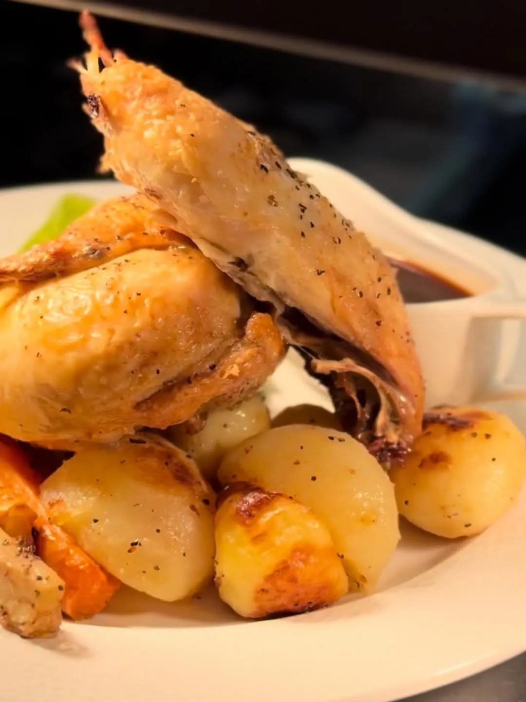 A professionally captured close-up of a roasted chicken dish, complete with golden-brown skin, roasted potatoes, and a gravy boat in the background. The image is well-composed and lit, with key elements in sharp focus, presenting a highly photo-realistic and appetizing view of the meal.