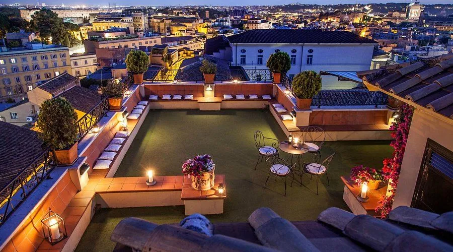 An elevated view of a hotel rooftop terrace at dusk or night, featuring comfortable seating arrangements, illuminated tables, and a panoramic city skyline in the background.