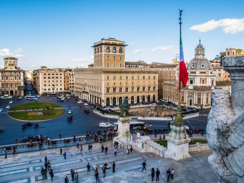 The image is well-composed with good natural lighting, showcasing a vibrant city scene with clear architectural details and an Italian flag. The colors are accurate and the image is sharp throughout, contributing to its strong photo-realistic quality. Key elements like the buildings and the flag are in clear focus. The scene, while busy with people and traffic typical of an urban environment, maintains an editorial feel.
