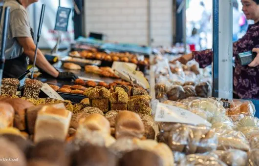 An editorial-style image showcasing a vibrant array of baked goods, including various breads and pastries, on display at a market stall or bakery. The image uses shallow depth of field to draw attention to the foreground and middle-ground items, highlighting their textures and freshness.