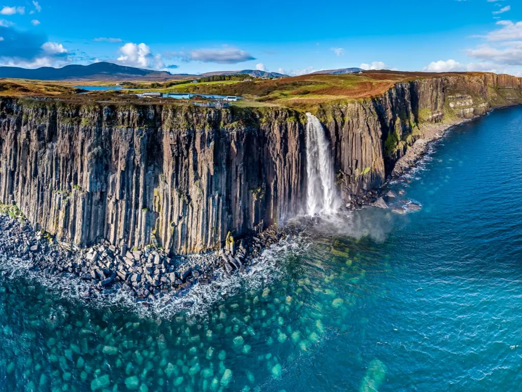An aerial view of a majestic waterfall cascading over dramatic basalt columns into the vibrant blue ocean. The composition captures the scale of the cliffs and the clarity of the water, with clear blue skies overhead and distant green hills.
