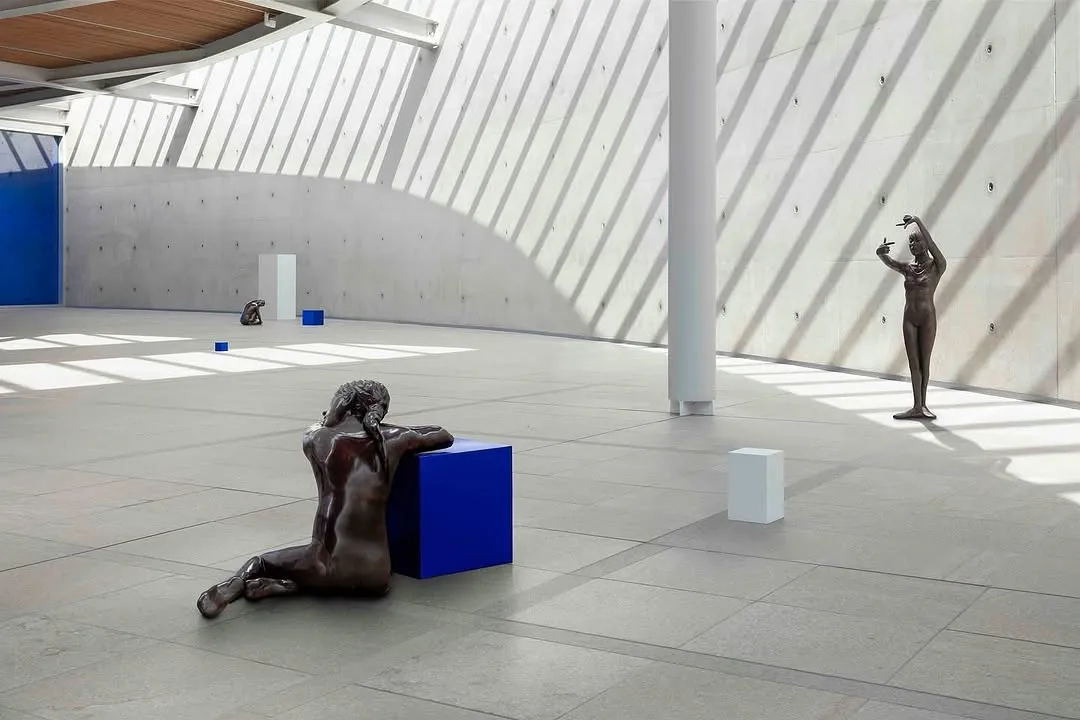 A well-composed and sharply focused photograph of an art installation featuring a bronze sculpture and blue cubes within a modern, naturally lit architectural space. The lighting creates striking shadow patterns, enhancing the depth and visual interest.