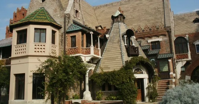 A detailed exterior view of an old building featuring a decorative balcony, a distinctive conical roof, and climbing ivy.