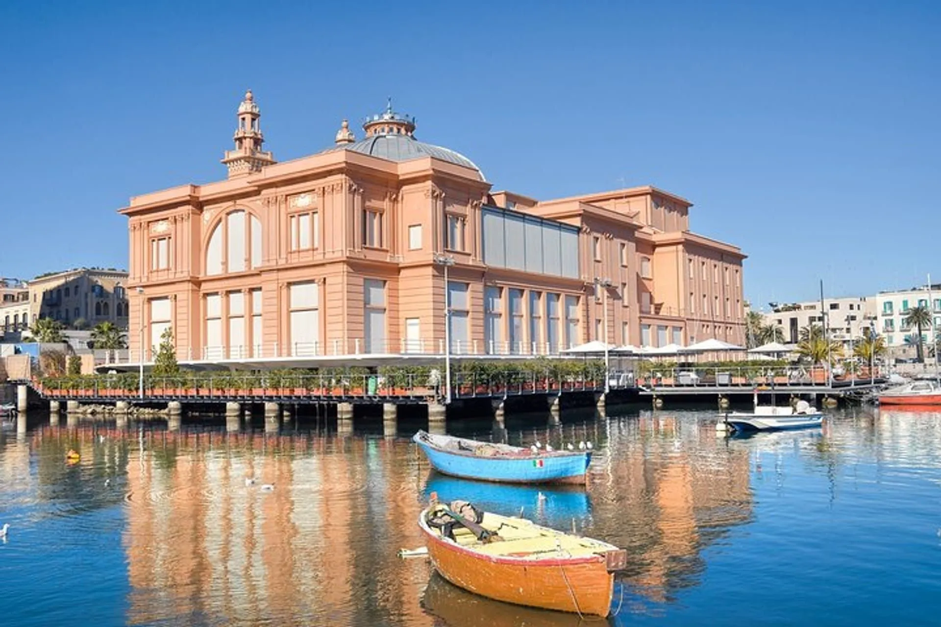 A vibrant architectural building with a distinctive dome and towers, situated next to a body of water, possibly a canal or harbor, where two colorful wooden boats are moored. The sky is clear blue, and reflections of the building and boats are visible in the water.