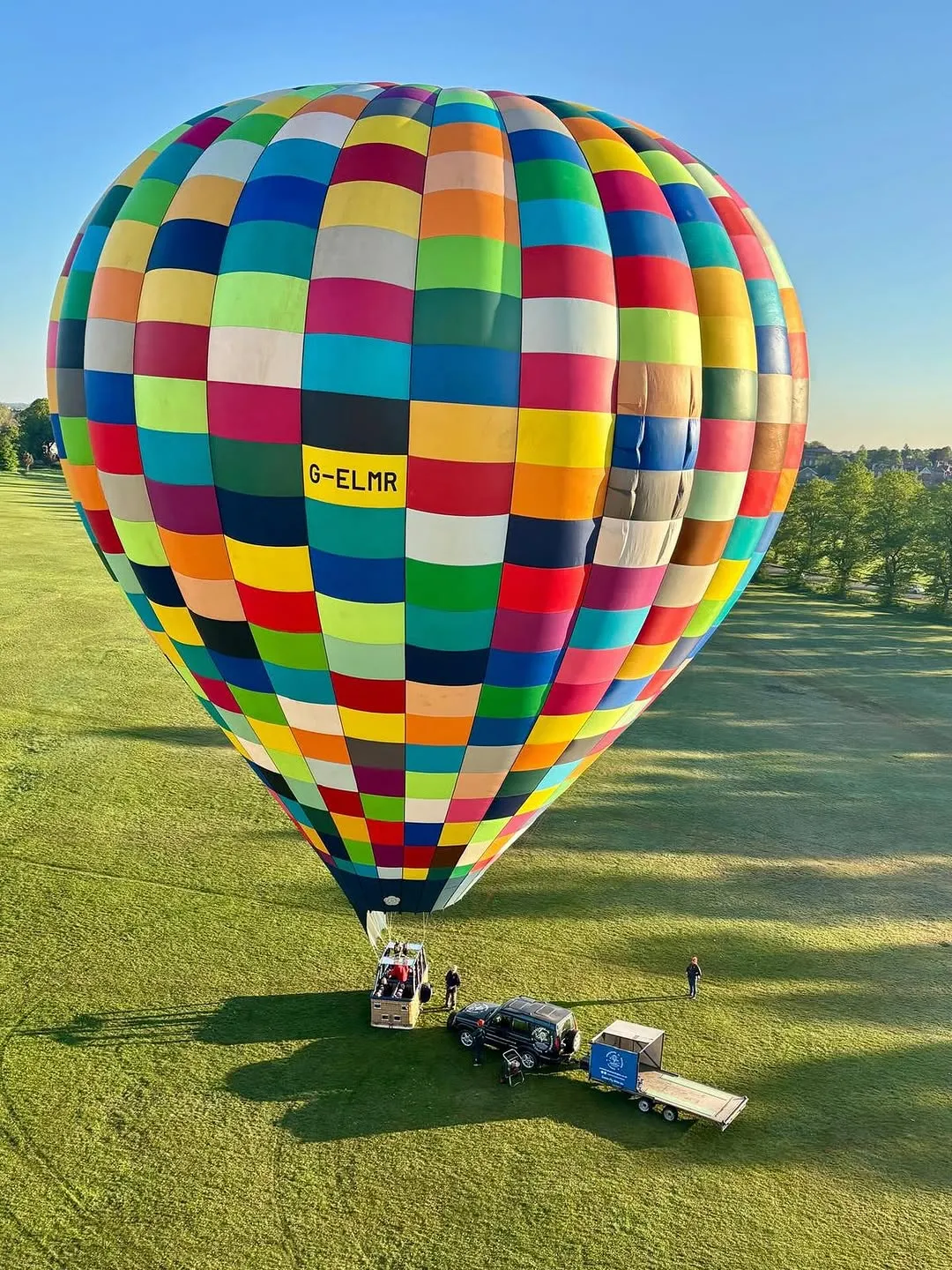 An aerial or elevated view of a large, vibrantly colored hot air balloon standing on a green grassy field under a clear blue sky, with a support vehicle, trailer, and a few people visible on the ground.
