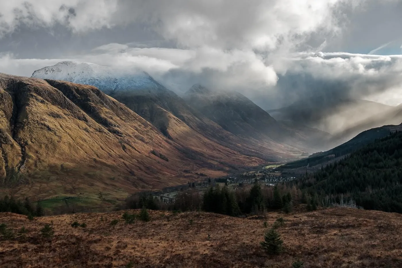 A dramatic wide shot of a mountainous valley featuring rugged brown and green slopes, partially obscured by low-hanging, moody clouds and mist. Patches of snow are visible on some of the higher peaks, and a small village with scattered trees is nestled deep within the valley floor.