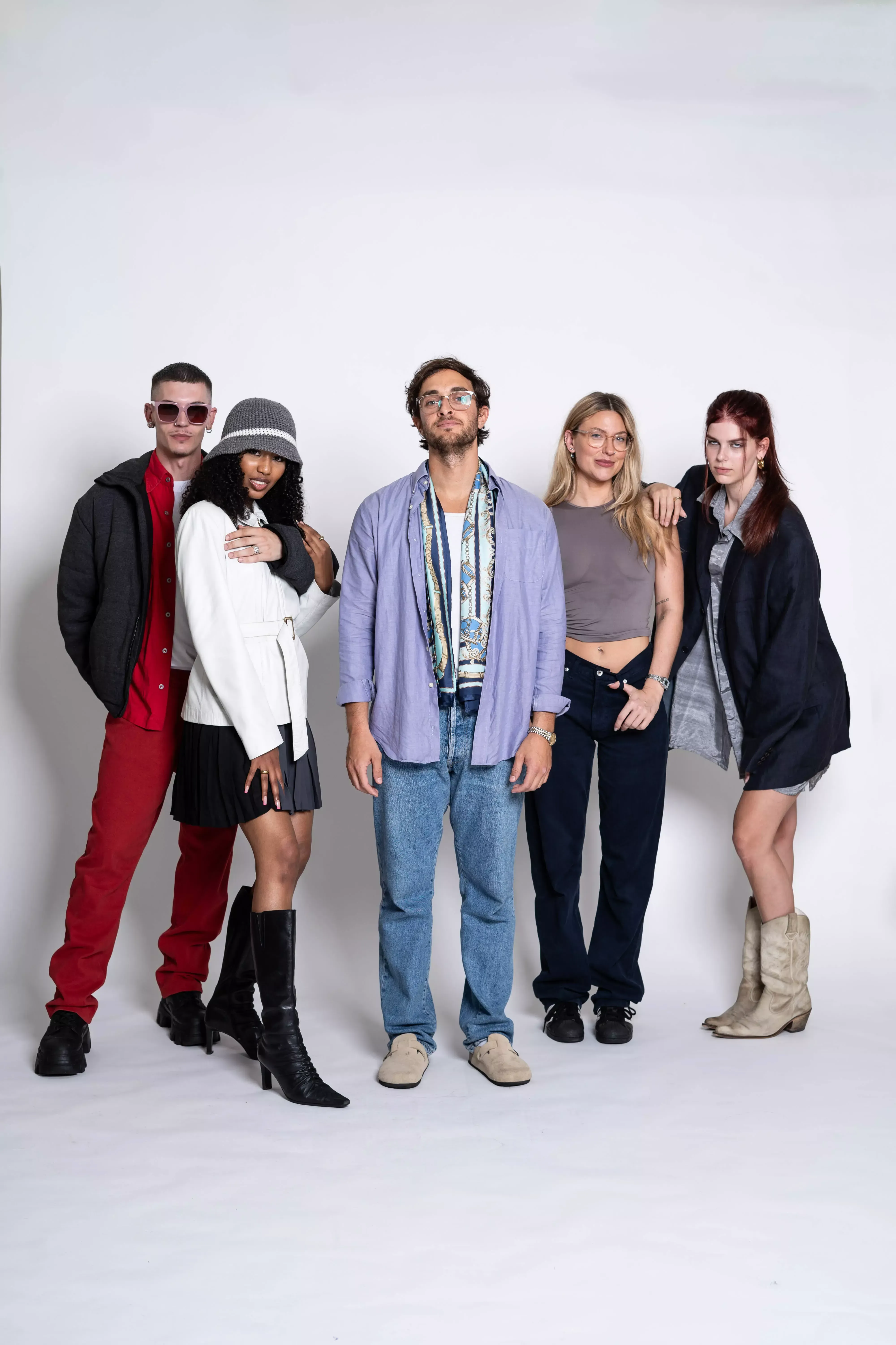 A group of five young adults, three women and two men, are standing against a plain white background, showcasing various fashion styles. They are dressed in a mix of casual and semi-casual attire, characteristic of an editorial photoshoot.