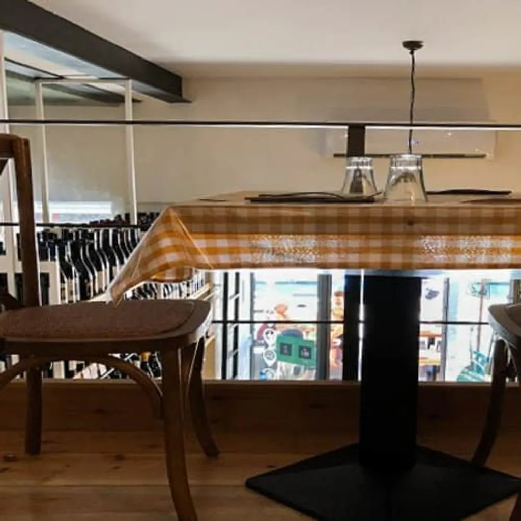 An interior shot of a restaurant dining area featuring a table set with an orange and white checkered tablecloth and two upside-down glasses. Two wooden chairs are pulled up to the table, and a black hanging light fixture is visible above. The background shows a railing and a view into another section of the establishment with what appears to be wine bottles.
