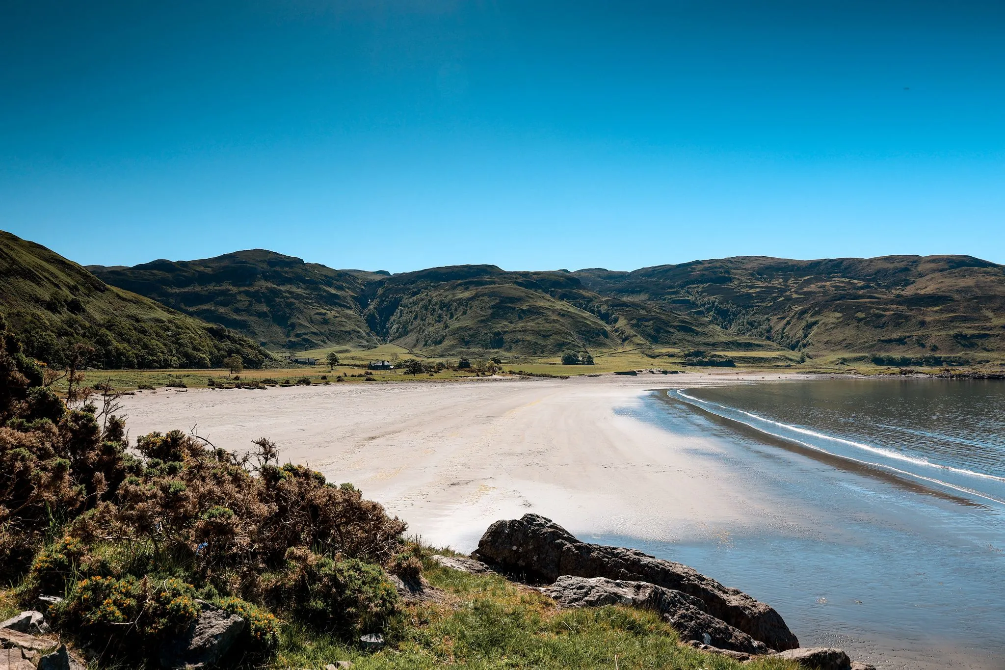 A beautiful, well-composed landscape image featuring a wide sandy beach meeting calm water, with rolling green mountains under a clear blue sky in the background. Foreground elements like rocks and low-lying bushes add depth to the scene.
