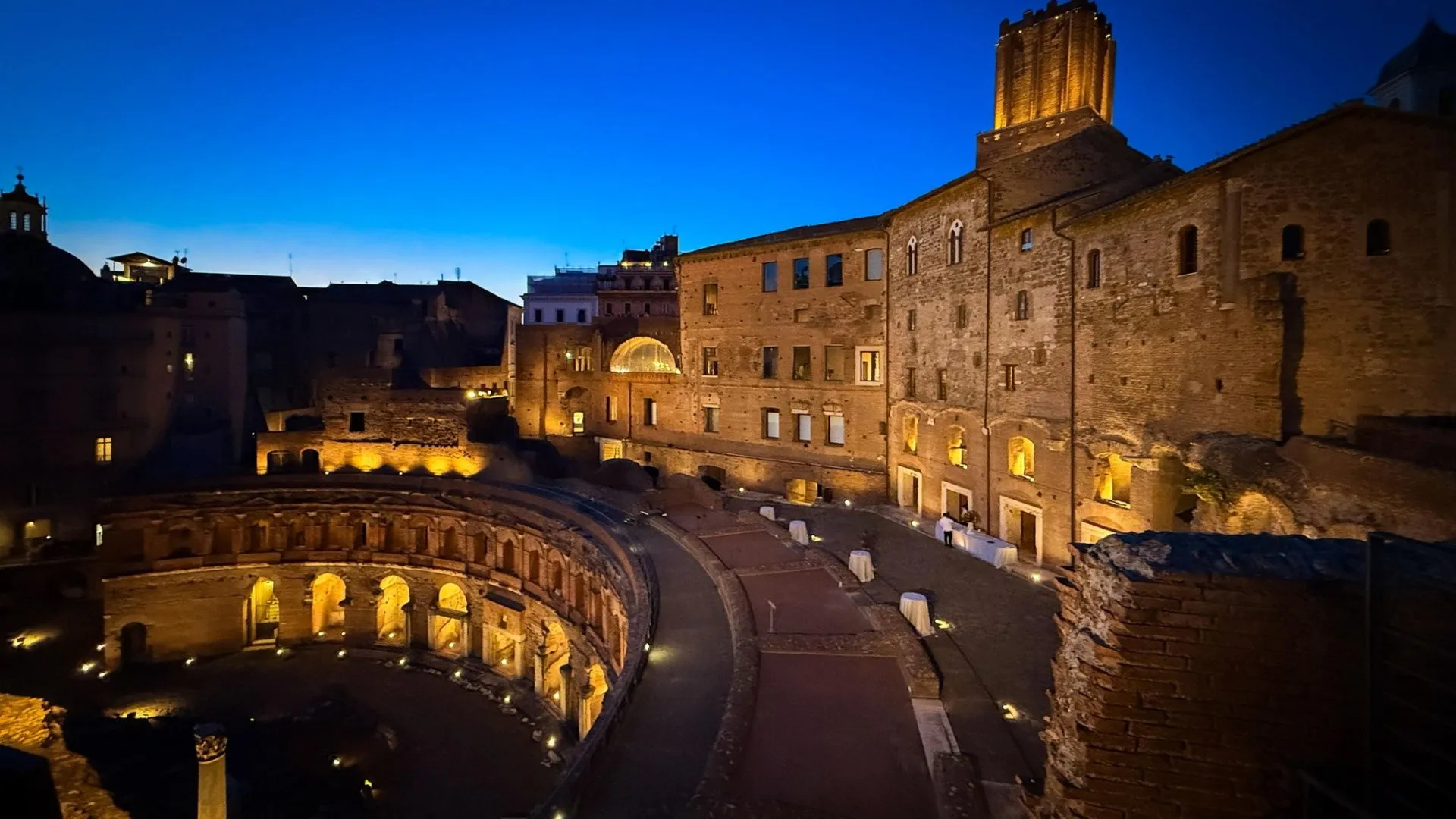 The image effectively captures the ancient Trajan's Market at dusk, showcasing its illuminated architecture against a deep blue sky. The composition is strong, drawing the eye through the curved structures. The lighting accentuates the textures and details of the historical buildings, contributing to a realistic and evocative scene. While generally sharp, some very dark areas could benefit from slightly more detail.