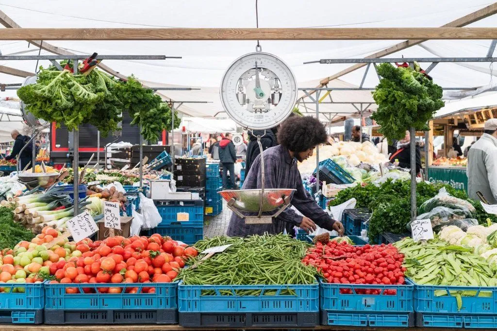 An editorial-style image capturing a vibrant outdoor market scene. The composition is strong, with a vendor interacting with fresh produce, prominently featuring crates of tomatoes and green beans in the foreground. A traditional weighing scale hangs above. The lighting is soft and even, accurately rendering the rich colors of the produce, and the image is sharp and photo-realistic, with key elements in focus. The busy market atmosphere is well-captured without being overly cluttered.