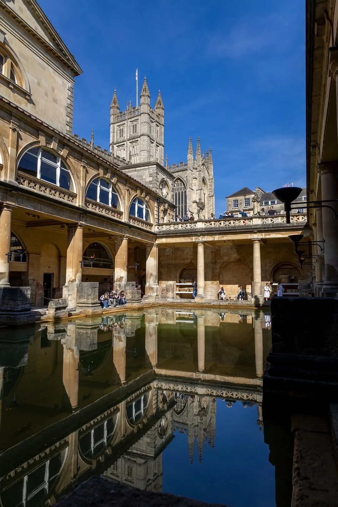A well-composed and sharp photograph of the Roman Baths, featuring the ancient architecture and its reflection in the water, with Bath Abbey prominently in the background. The image is well-lit with natural sunlight, showcasing accurate colors and high photo-realism. Key elements are in sharp focus, providing a clear view of the historical site. The presence of people does not detract from the scene.
