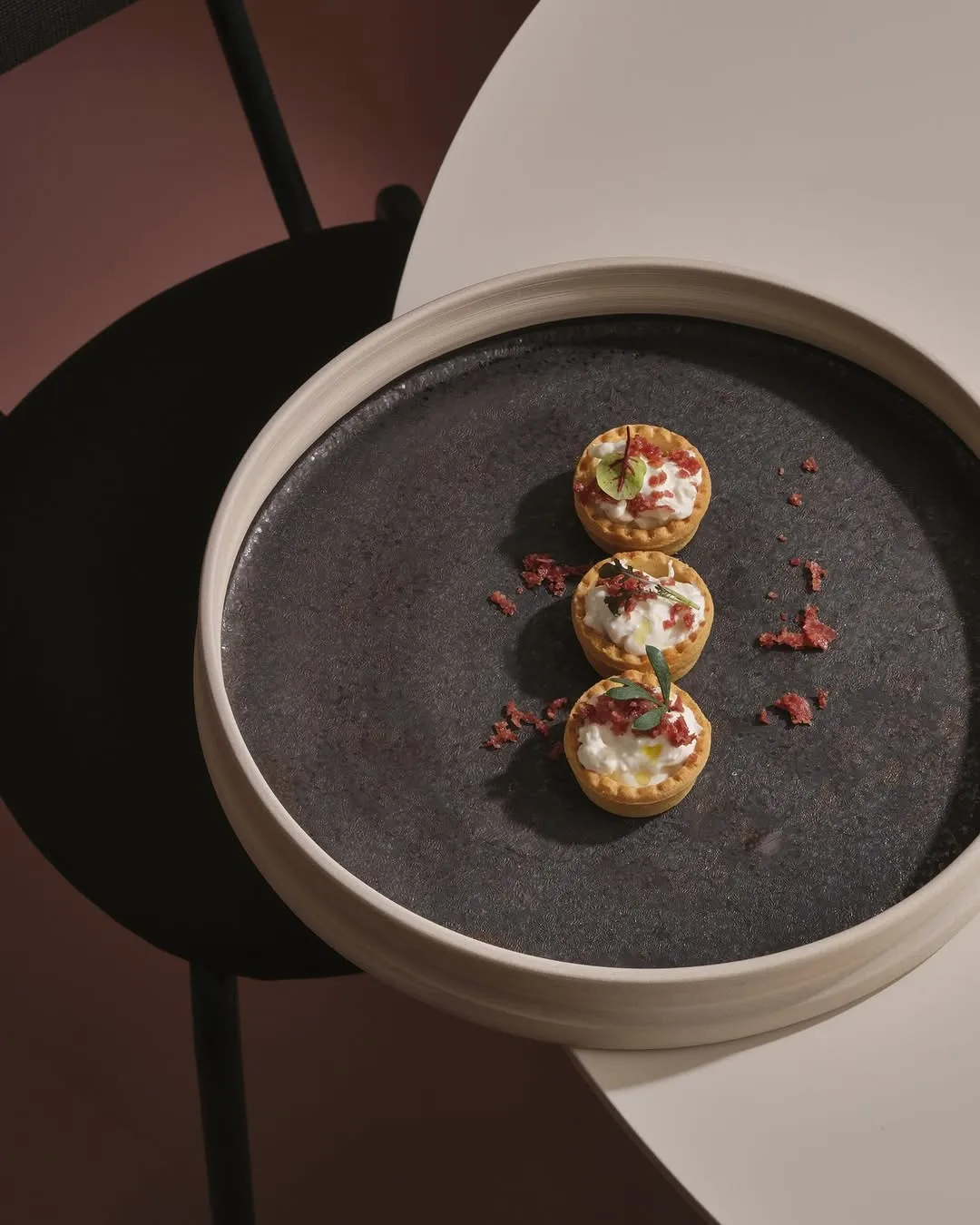 A professionally taken, high-quality image showcasing three small, savory tartlets meticulously arranged on a dark, textured ceramic plate. The background is simple and uncluttered, featuring a blend of dark and lighter tones with artistic shadows that add depth. The tartlets are golden-brown with creamy white filling, garnished with red crumbs and small green herbs, suggesting a gourmet appetizer or canapé.
