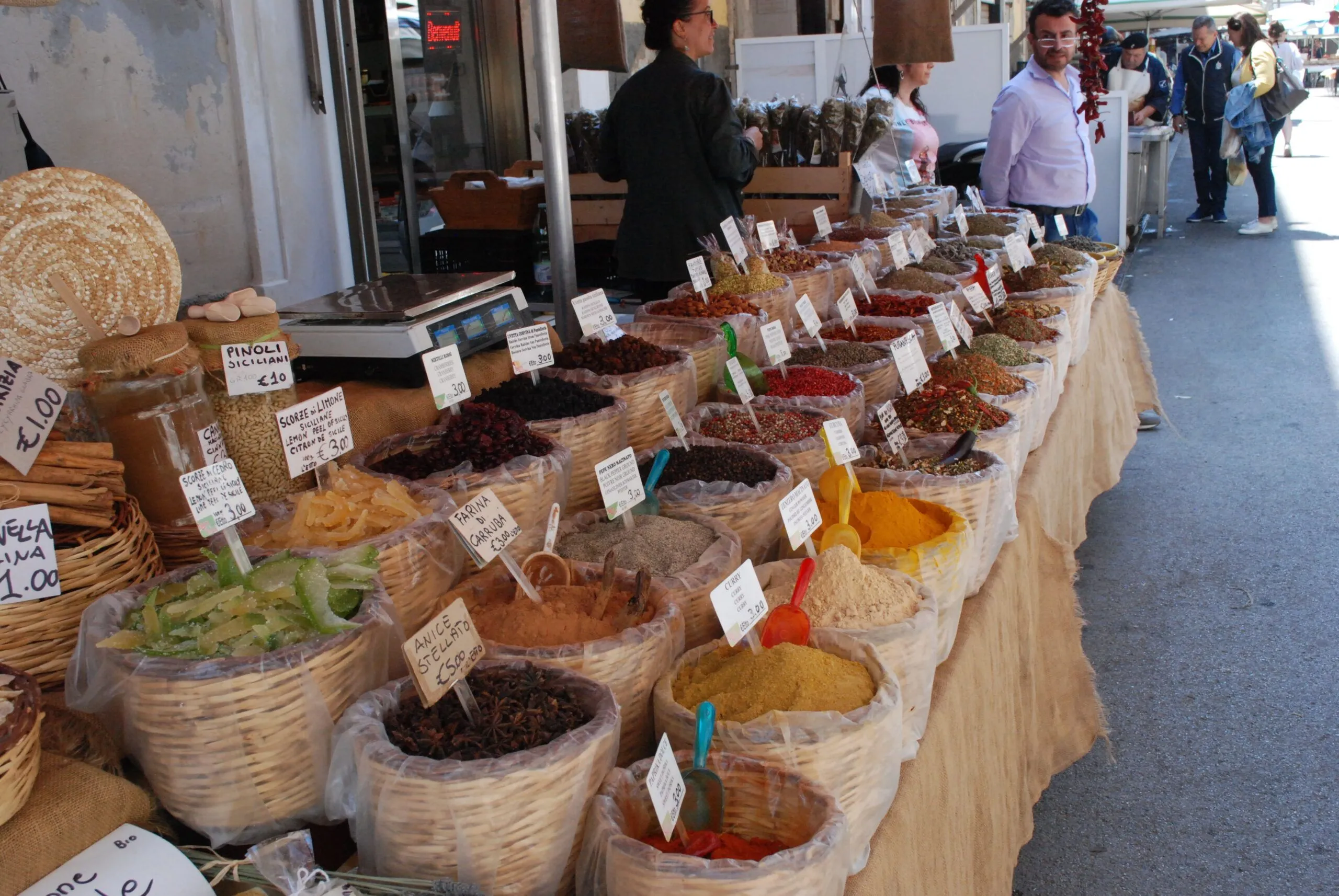 An editorial-style image capturing a vibrant market stall filled with various spices and dried goods in woven baskets. Price tags are visible on many items, and people are in the background, adding to the market atmosphere.
