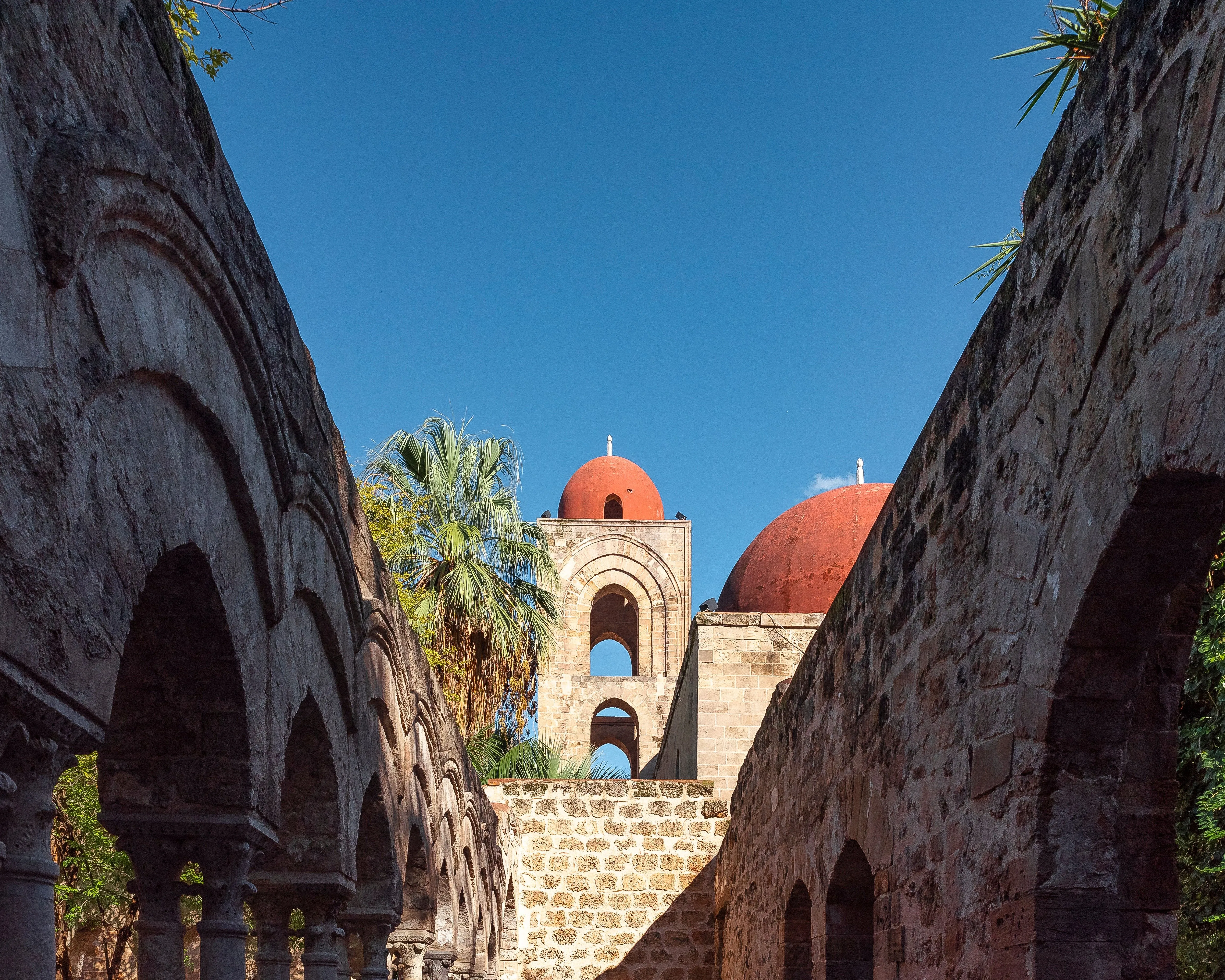 An impressive architectural shot featuring historic stone buildings with prominent red domes set against a vivid blue sky. The composition expertly uses natural framing from arches and walls to draw the eye towards the central structures.