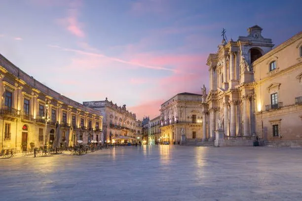 An evocative image of a historic European city square at twilight, showcasing impressive classical architecture. The sky is painted with soft hues of pink and blue, creating a serene and picturesque atmosphere. Warm lights from the buildings illuminate the facades and reflect gently on the stone ground of the square.