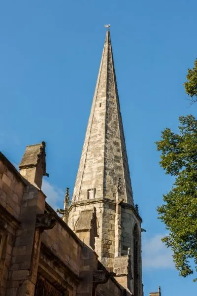 A striking editorial image showcasing a tall, weathered stone church spire set against a vibrant blue sky, with architectural details of an adjacent building and green tree foliage framing the scene. The lighting is bright and clear, highlighting the intricate texture of the stone.