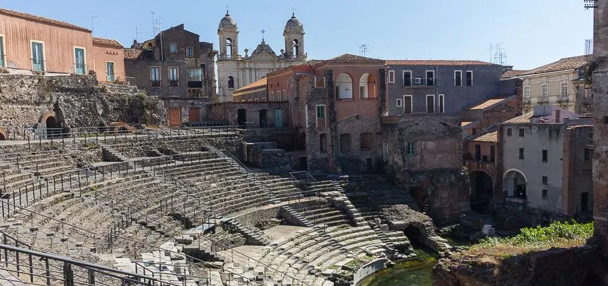 An editorial image showcasing ancient Roman or Greek theatre ruins with surrounding traditional buildings and a church in the background. The image provides a good perspective of the historical site integrated into a city environment.