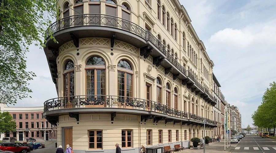 An editorial-style photograph showcasing the architectural beauty of a historic building. The image features a cream-colored facade with intricate iron balconies and arched windows, captured under natural lighting.