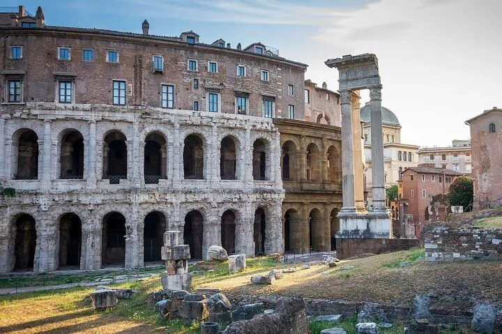 An editorial-style image capturing the ancient Theatre of Marcellus in Rome, bathed in natural sunlight. The composition highlights the historical stone structure with its arches and partially preserved columns, contrasting with a building behind it. The foreground shows ruins and overgrown grass, adding to the sense of antiquity.