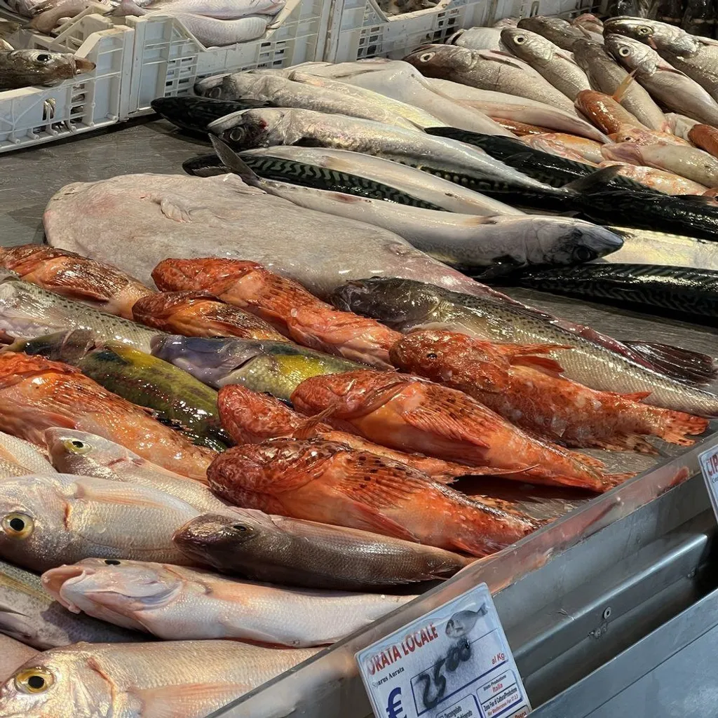 A close-up, editorial-style image showing a variety of fresh fish, including red scorpionfish and mackerel, displayed on a counter at a market. A price tag for "ORATA LOCALE" is visible in the foreground.