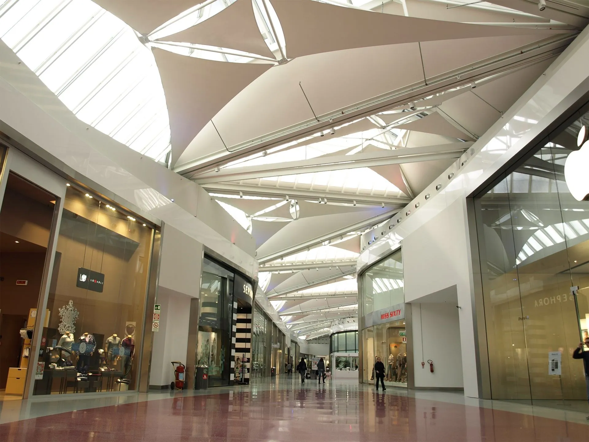 An interior view of a modern shopping mall featuring a distinctive architectural ceiling with skylights, long hallways, and various storefronts.