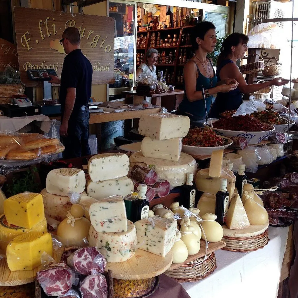 An editorial image showcasing a vibrant market stall filled with various cheeses, cured meats, and other specialty food items. The foreground is laden with different types of cheese wheels, blocks, and cured sausage, beautifully arranged on wooden and woven surfaces. In the background, vendors interact with customers, adding to the bustling market atmosphere. The scene captures the essence of a traditional food market.