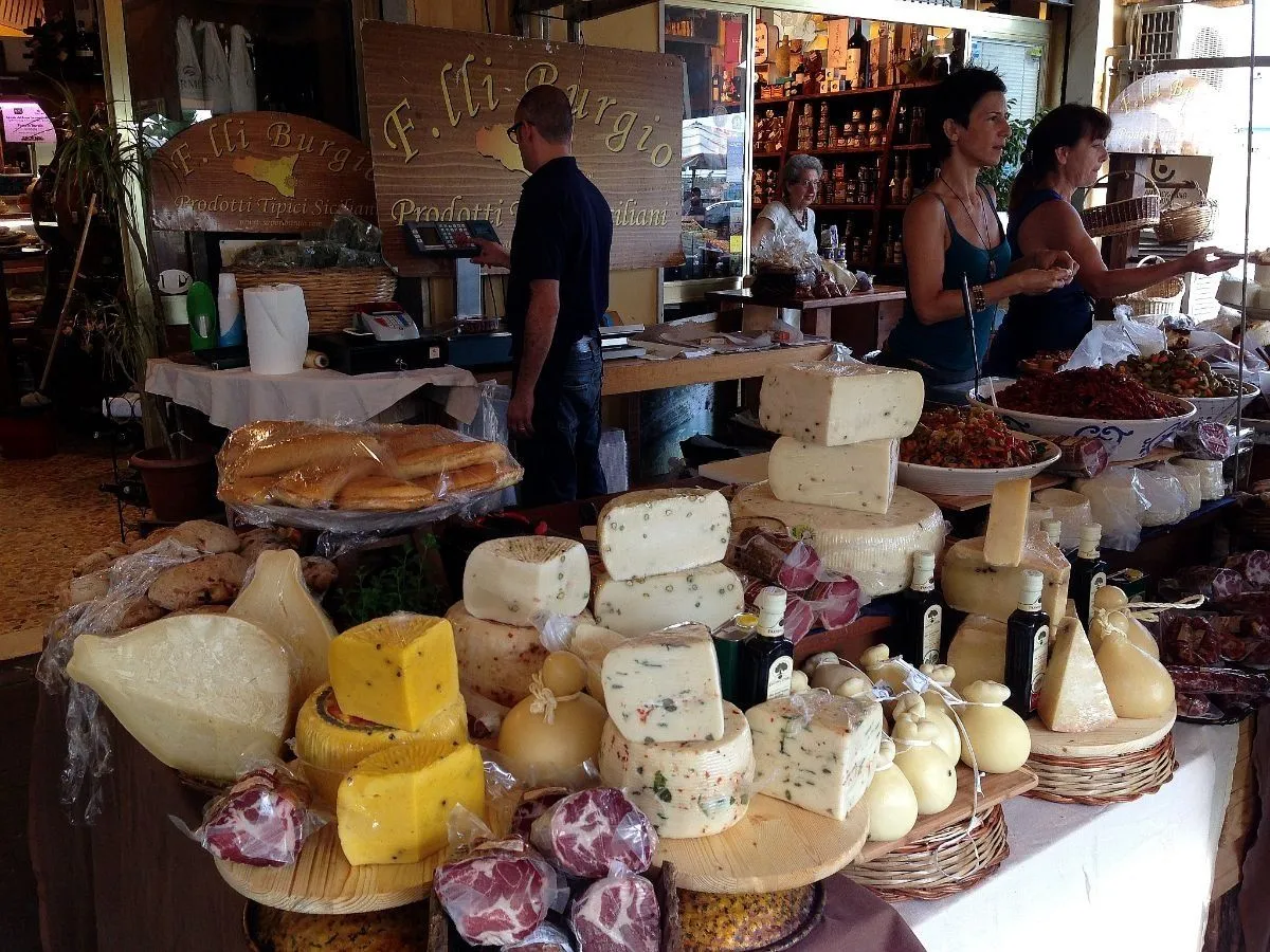An editorial image showcasing a vibrant market stall filled with various cheeses, cured meats, and other specialty food items. The foreground is laden with different types of cheese wheels, blocks, and cured sausage, beautifully arranged on wooden and woven surfaces. In the background, vendors interact with customers, adding to the bustling market atmosphere. The scene captures the essence of a traditional food market.