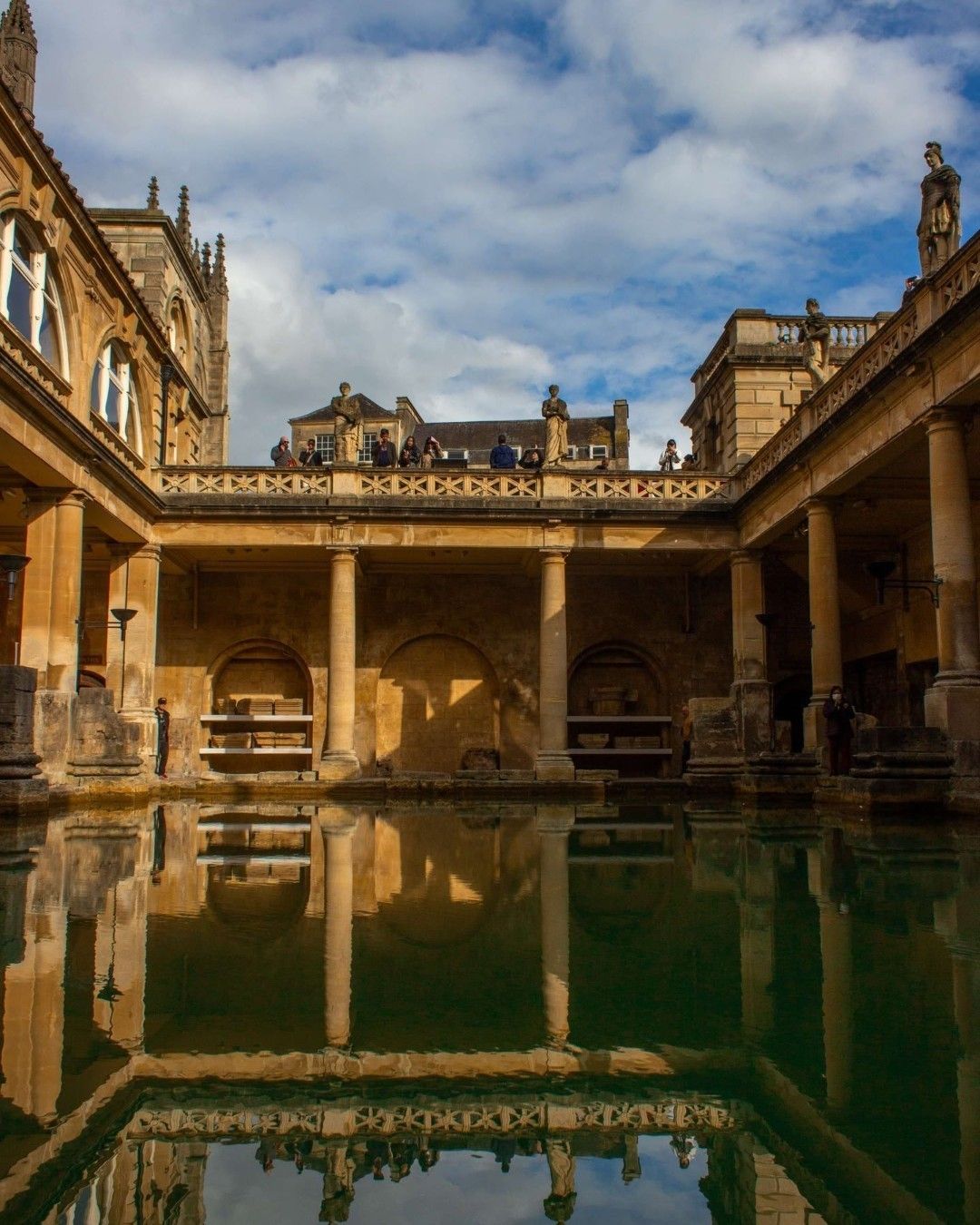This image is a professionally taken photograph of the Roman Baths in Bath, England. The composition is excellent, showcasing the symmetry and reflections in the water. The lighting is well-balanced, highlighting the architectural details without harsh shadows. The image is sharp, with clear details visible throughout. Colors are accurate, reflecting the natural tones of the stone. The photo is highly photorealistic and all key elements (the architecture and reflections) are in sharp focus. There are no unwanted elements present.