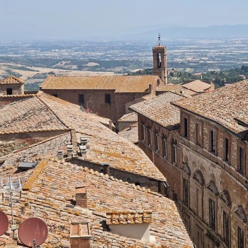 An aerial view of an old European city, showcasing terracotta rooftops and historic buildings under a clear sky, with a distant bell tower and hazy landscape.
