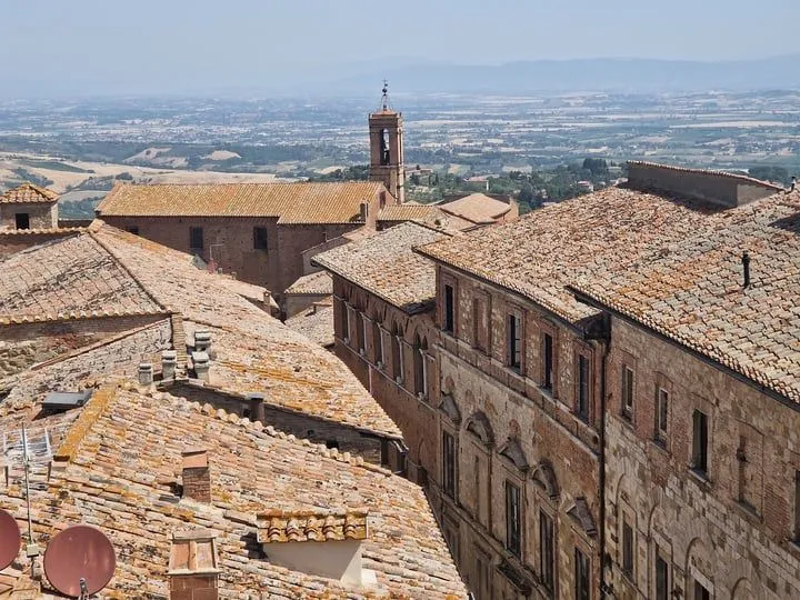 An aerial view of an old European city, showcasing terracotta rooftops and historic buildings under a clear sky, with a distant bell tower and hazy landscape.