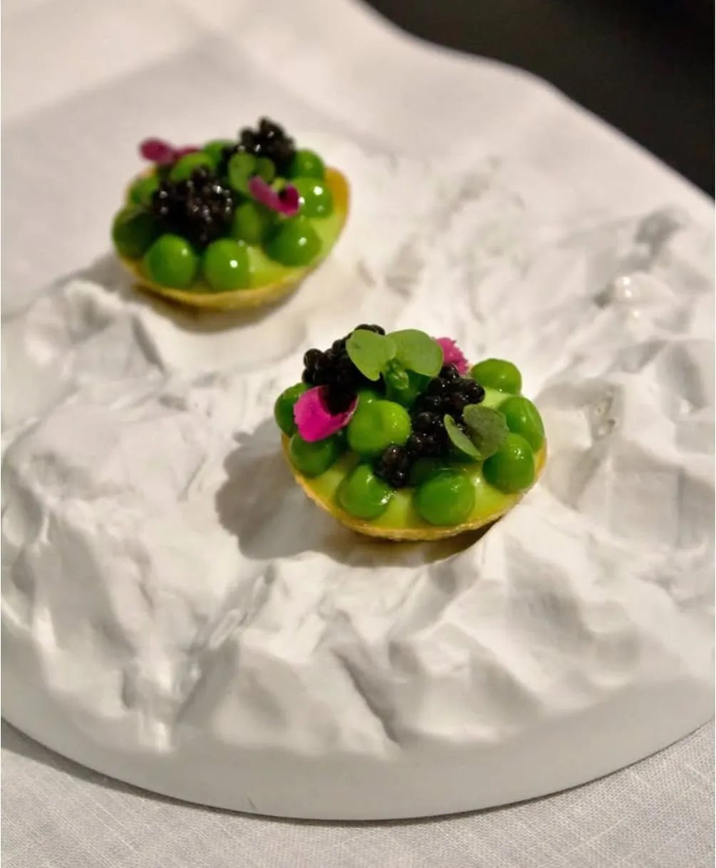 The image features two exquisitely presented food items on a unique, textured white plate. The lighting is soft and even, highlighting the vibrant green of the peas, the dark sheen of the caviar, and the delicate pink petals. The main subject in the foreground is sharp and detailed, contributing to a highly photo-realistic and appetizing appearance.