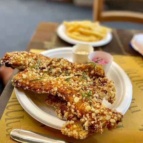 A close-up, editorial-style shot of golden-brown fried food, likely chicken or fish, topped with a crispy, textured coating and green herbs, served on a white plate. Two small cups containing sauces are next to the main dish, and a blurred plate of french fries is visible in the background, set on a patterned table.