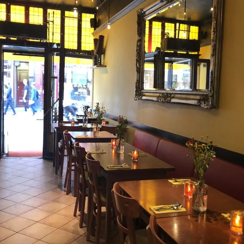 A warm and inviting restaurant interior featuring rows of wooden tables set with cutlery and candles, red booth seating, and a decorative mirror, looking towards a bright street entrance.