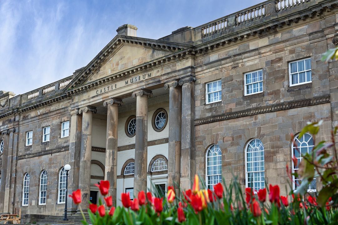 The image showcases the exterior of a castle museum, capturing its architectural details and surrounding greenery. The composition is well-balanced, leading the viewer's eye towards the imposing entrance. The lighting is excellent, highlighting the stonework and textures of the building. The image is sharp and color-accurate, showcasing the details of the building and the vibrant hues of the tulips. The key elements, including the building facade and the flowers, are in focus. The image effectively conveys the grandeur and historical significance of the castle museum. The only minor drawback is the presence of text on the building, which is considered an unwanted element. However, the text is subtle and does not detract significantly from the overall aesthetic.