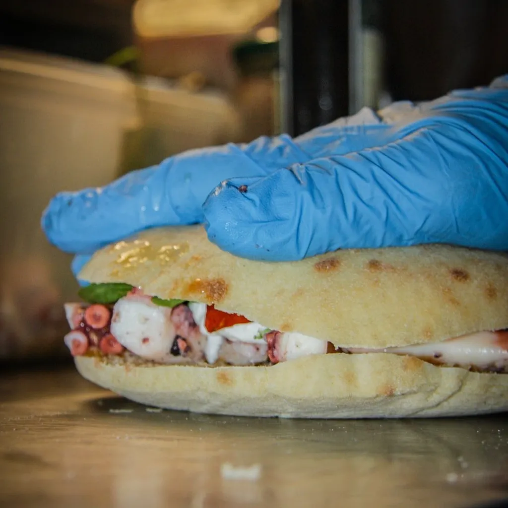 A professionally taken close-up image of a person wearing a blue disposable glove holding a savory sandwich or flatbread filled with octopus, tomatoes, and greens. The image is sharp, highlighting the textures of the bread and the seafood, and the colors are vibrant and accurate. The background is softly blurred, focusing attention on the food preparation.