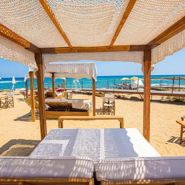 An editorial style image showcasing a serene beach resort setup. The view is from underneath a wooden cabana with a white mesh canopy, looking out onto a sandy beach furnished with loungers and other cabanas. In the background, the clear blue ocean meets the horizon under a bright sky, with a wooden walkway extending near the water.