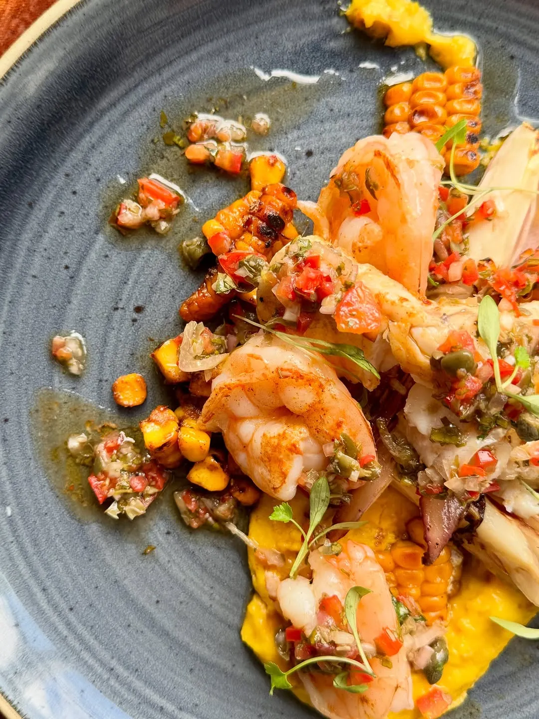 The image features a vibrant and well-composed close-up of a seafood dish, likely shrimp with a fresh salsa and grilled corn. The lighting is excellent, evenly illuminating the food and highlighting its textures and rich colors. Key elements, such as the shrimp and the colorful salsa, are sharp and in focus, contributing to a highly photo-realistic and appetizing presentation. The colors appear accurate and natural.