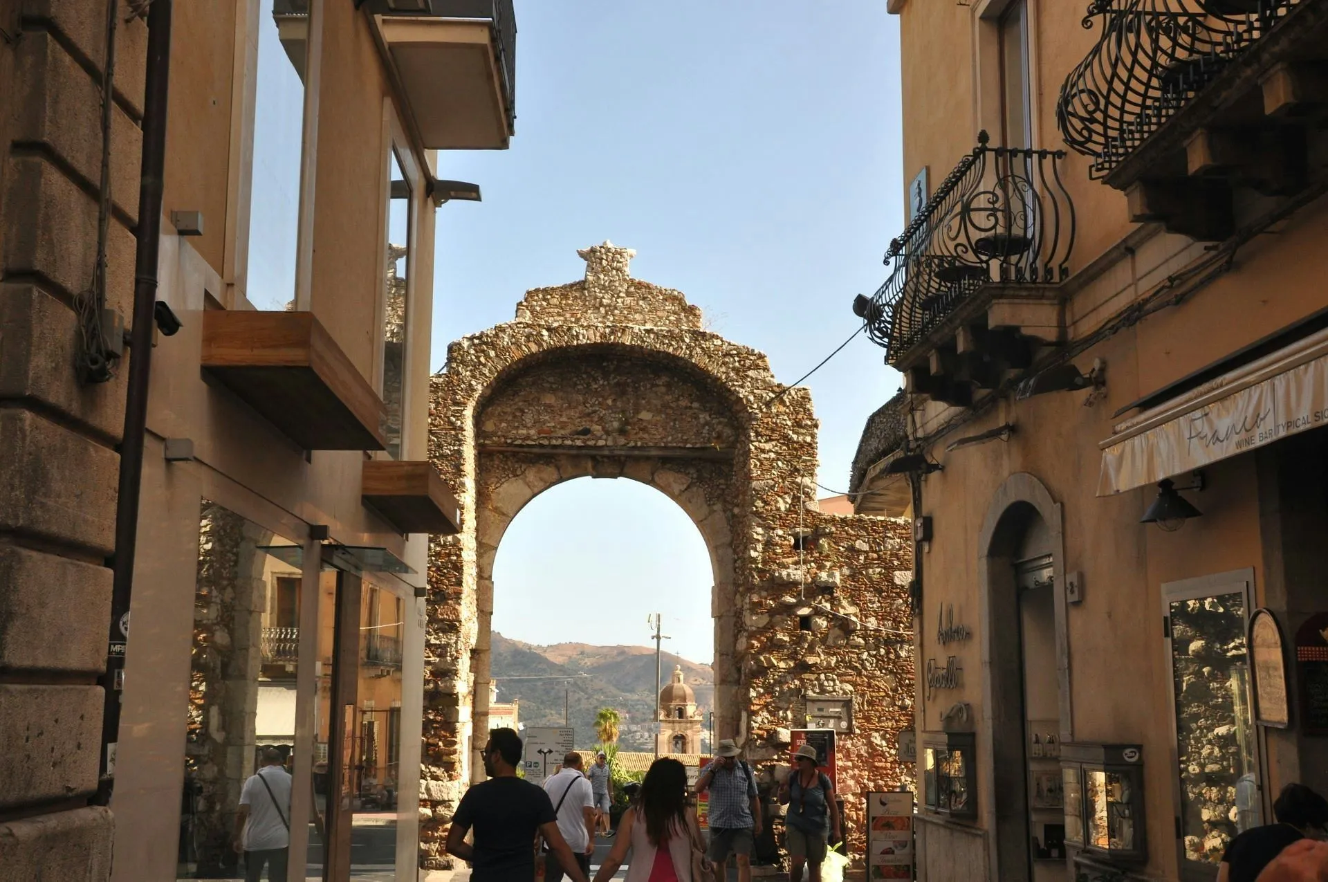 An editorial-style image capturing a street scene in a historic town, featuring an ancient stone archway framing a view of distant mountains and a church dome, with people walking through the street.