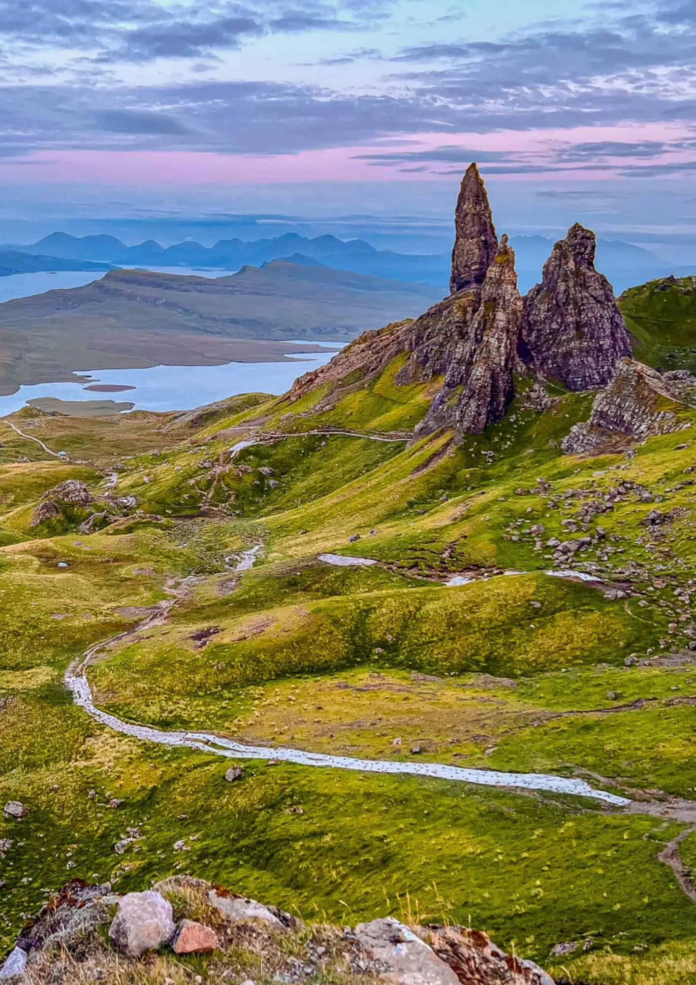 An expansive aerial view of the Old Man of Storr rock formations on the Isle of Skye, Scotland. The image captures a dramatic green landscape with a winding path leading towards a distant loch and mountains under a soft, colorful sky.