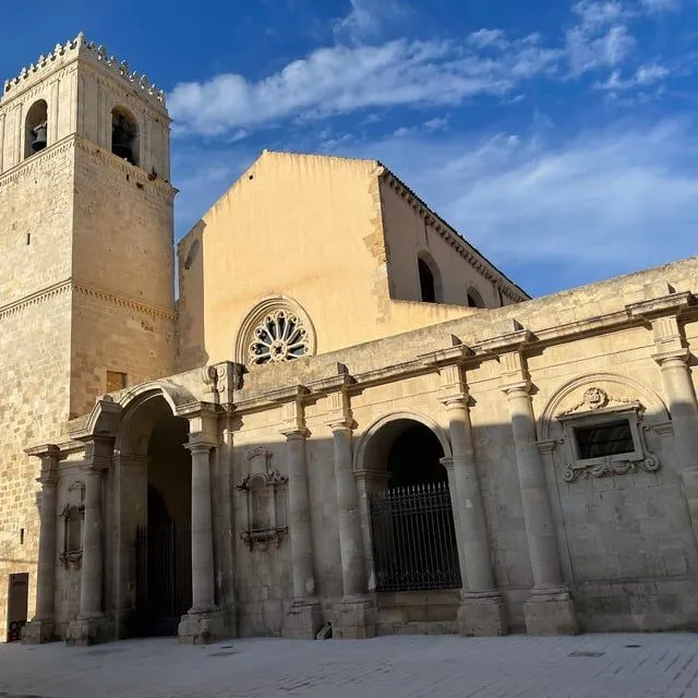 An architectural photograph of a historic building, likely a church or cathedral, featuring a bell tower and a rose window, set against a blue sky.