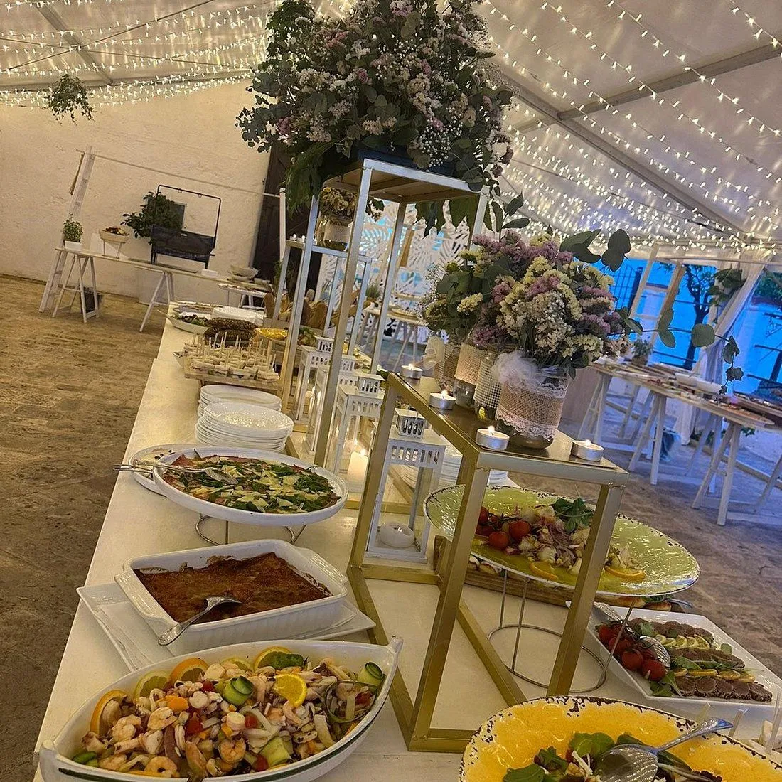 The image effectively captures a buffet spread at an event. Its strengths include good sharpness, realistic appearance, and most key elements being in focus. The composition is generally good, highlighting the food and decorations. However, the lighting, primarily from ambient string lights, is somewhat uneven and could be brighter, leading to some areas appearing a bit dim.