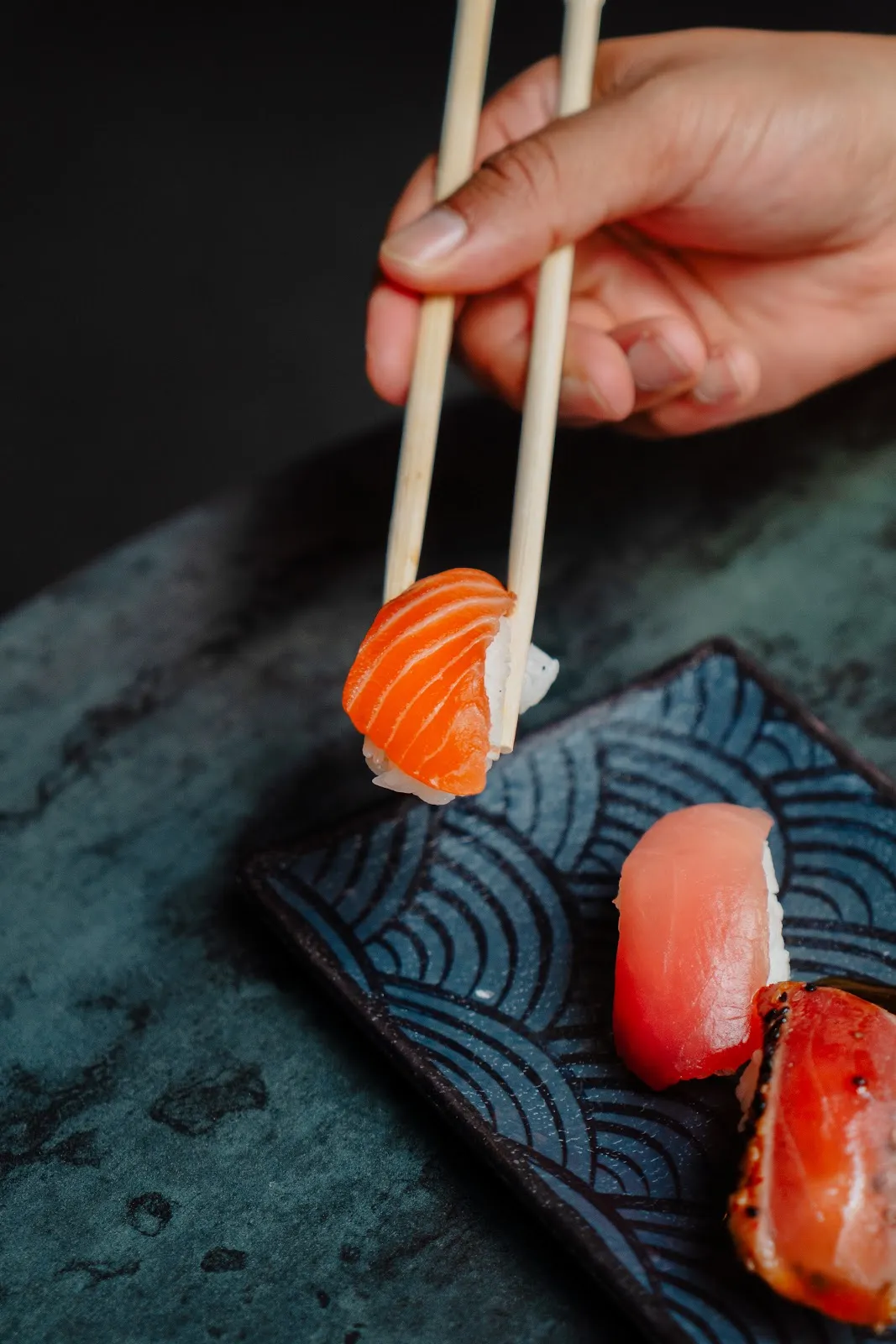 The image showcases a well-composed and sharply focused piece of salmon nigiri being held by chopsticks, creating a dynamic and appealing shot. The lighting effectively highlights the vibrant colors and fresh textures of the sushi, which appear highly photo-realistic and color-accurate. The key elements, particularly the sushi in the foreground, are perfectly in focus, while other elements provide context without distracting from the main subject.