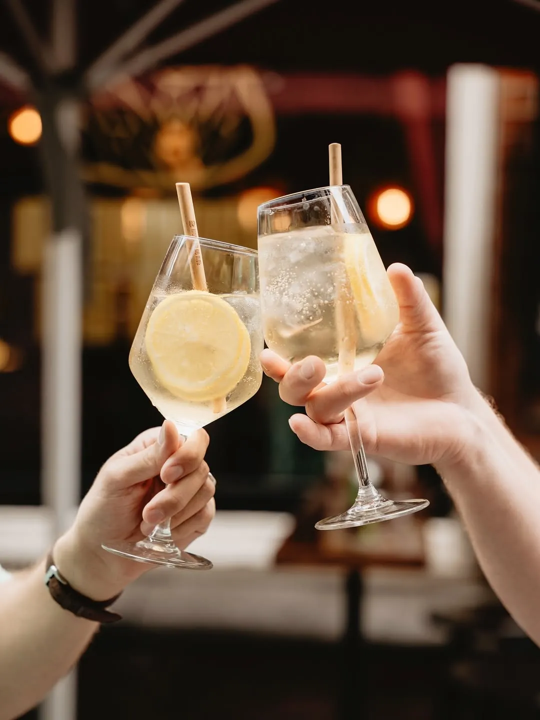 Two hands clink glasses filled with a light-colored, sparkling beverage, garnished with a lemon slice and a straw, against a softly blurred background with bokeh lights.