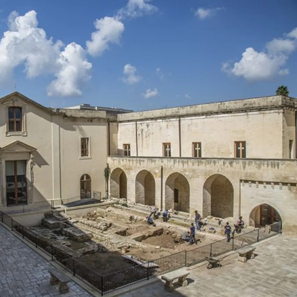 An outdoor shot of a historical courtyard featuring stone buildings and an active archaeological excavation site under a blue sky with white clouds.