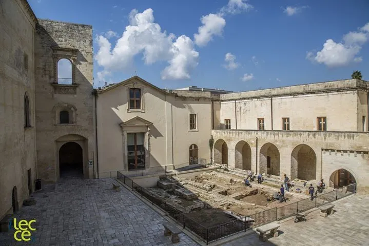 An outdoor shot of a historical courtyard featuring stone buildings and an active archaeological excavation site under a blue sky with white clouds.