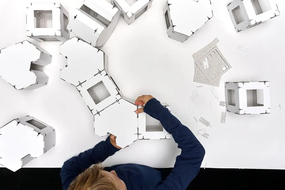 An overhead view of a person, likely a child, with blonde hair and a blue long-sleeved shirt, engaged in assembling white modular cardboard pieces on a clean white surface. Their hands are actively connecting the pieces, with other pre-assembled or unpunched components scattered around. The image focuses on the activity and the construction process.