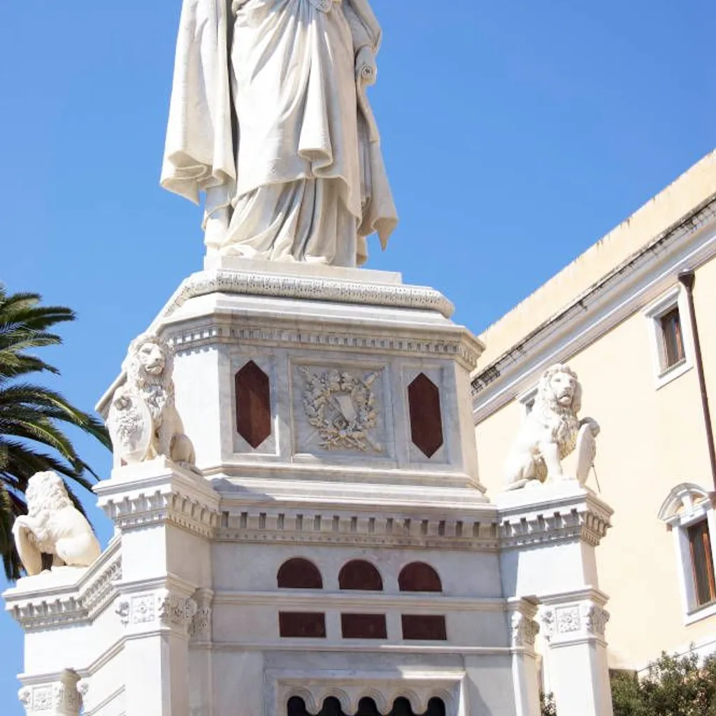The image features a large, white marble statue, likely a monument or landmark, set against a bright blue sky. The statue is well-lit by natural sunlight, showcasing intricate details and textures. The base of the statue is adorned with lion sculptures.