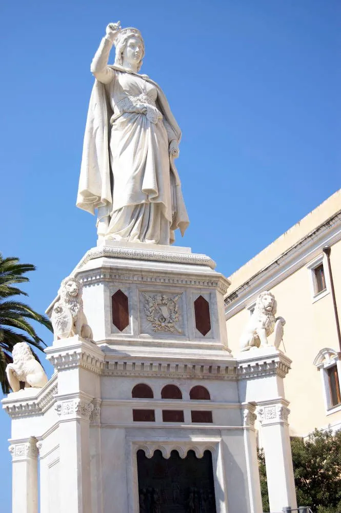 The image features a large, white marble statue, likely a monument or landmark, set against a bright blue sky. The statue is well-lit by natural sunlight, showcasing intricate details and textures. The base of the statue is adorned with lion sculptures.
