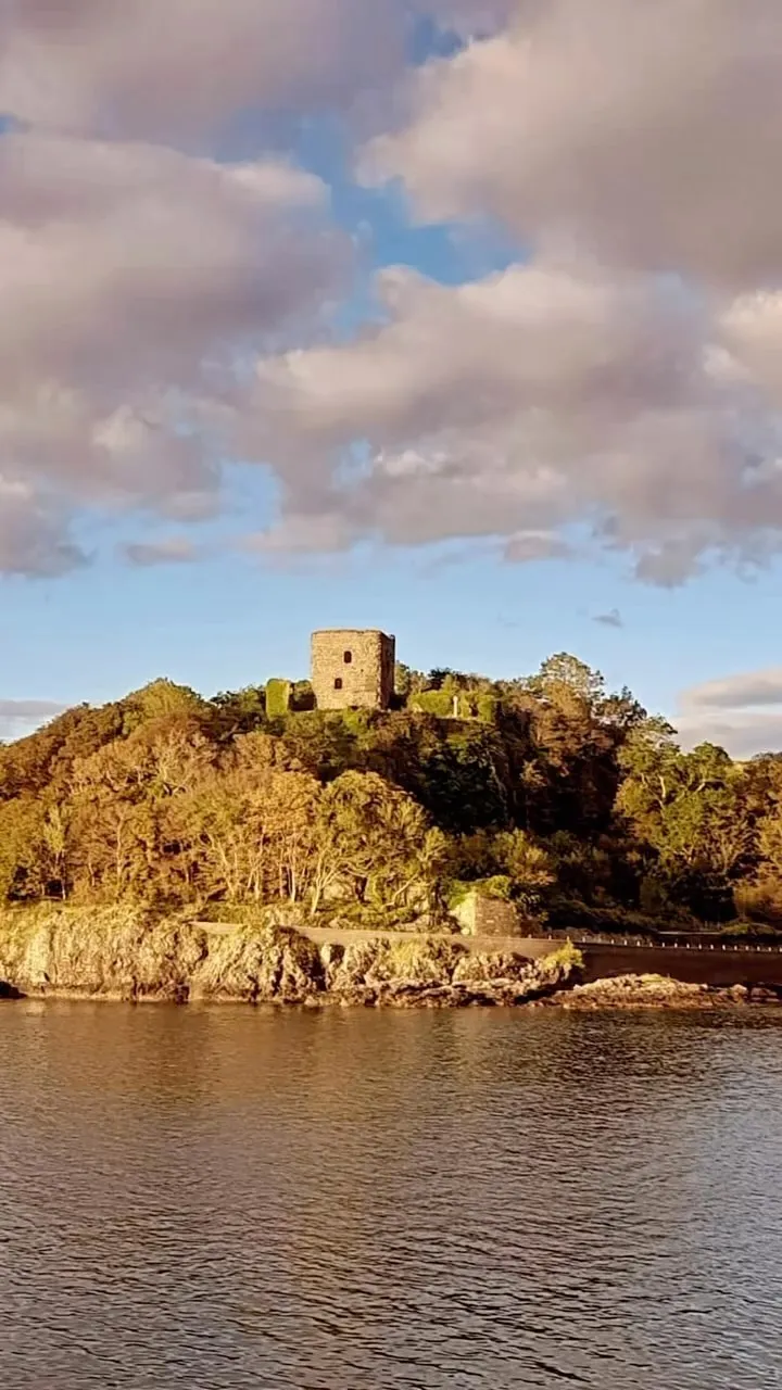 A stone castle stands prominently on a tree-covered hill, with a body of water in the foreground and a partly cloudy sky above.